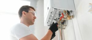 Tecnico idraulico con occhiali protettivi e guanti mentre esegue la riparazione o l'installazione di una caldaia a muro bianca.